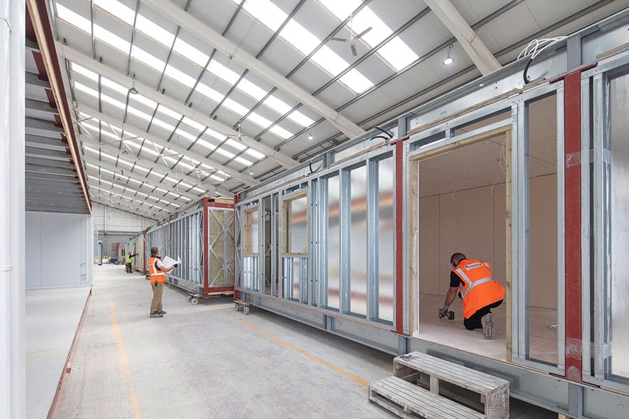 Inside Premier Modular's factory, modular units being worked on by two of our construction workers wearing orange high-vis vests.