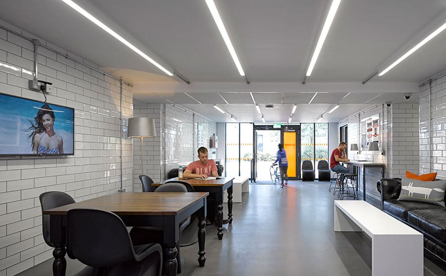 Communal area in modular accommodation building. TV screen on the wall. Tables and chairs, as well as benches.