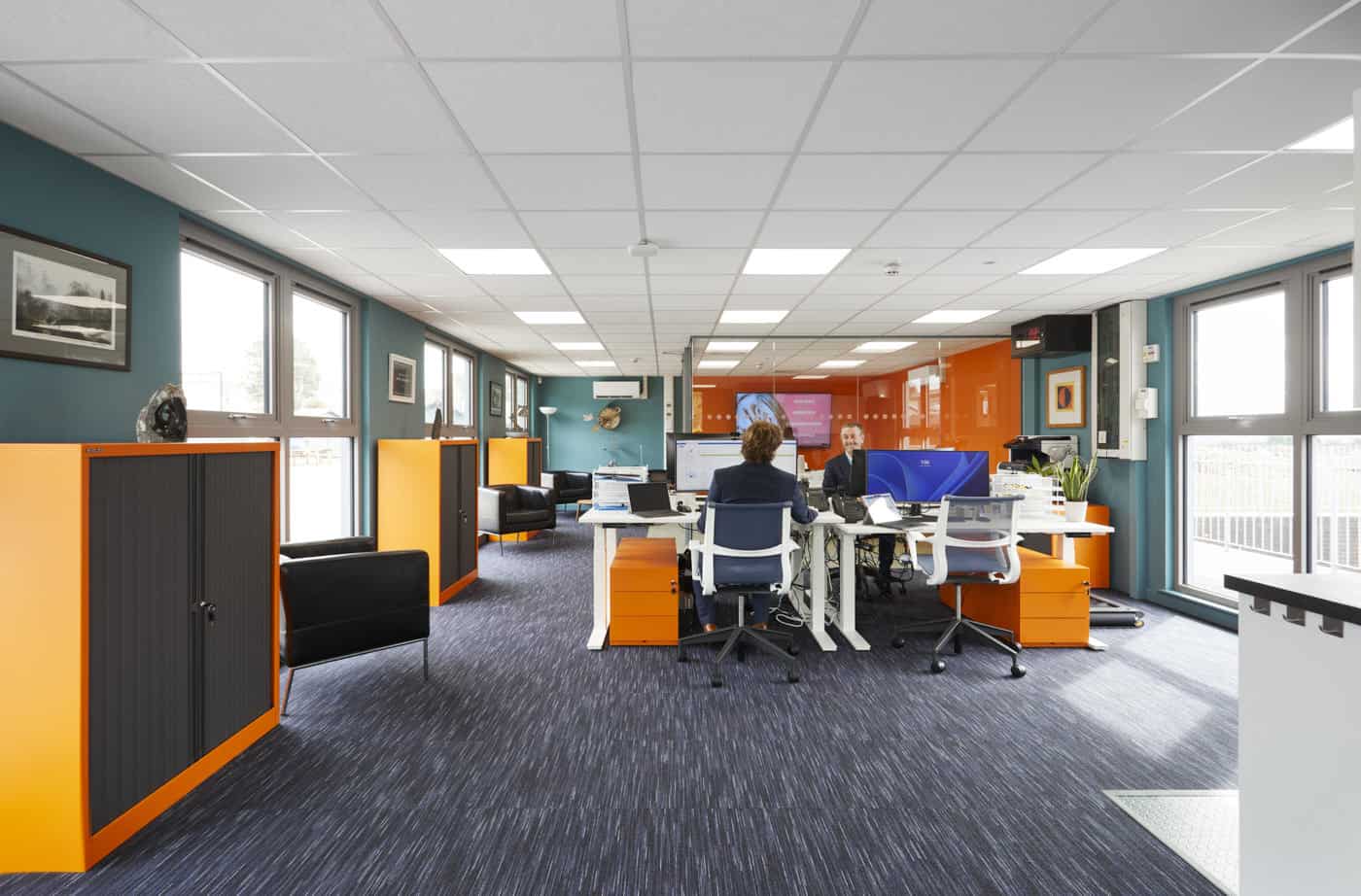 Inside a modular office facility. 2 people sat at computers on their desks in the centre of the room. colour fun blue and orange walls. Orange cabinets. Black arm chairs near the windows.