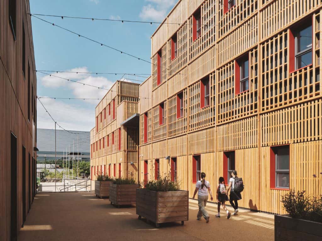 A family walking on the campus of The Paper yard, temporary modular structure. Multiple large buildings, with red window panes and wooden panelling on the walls. 