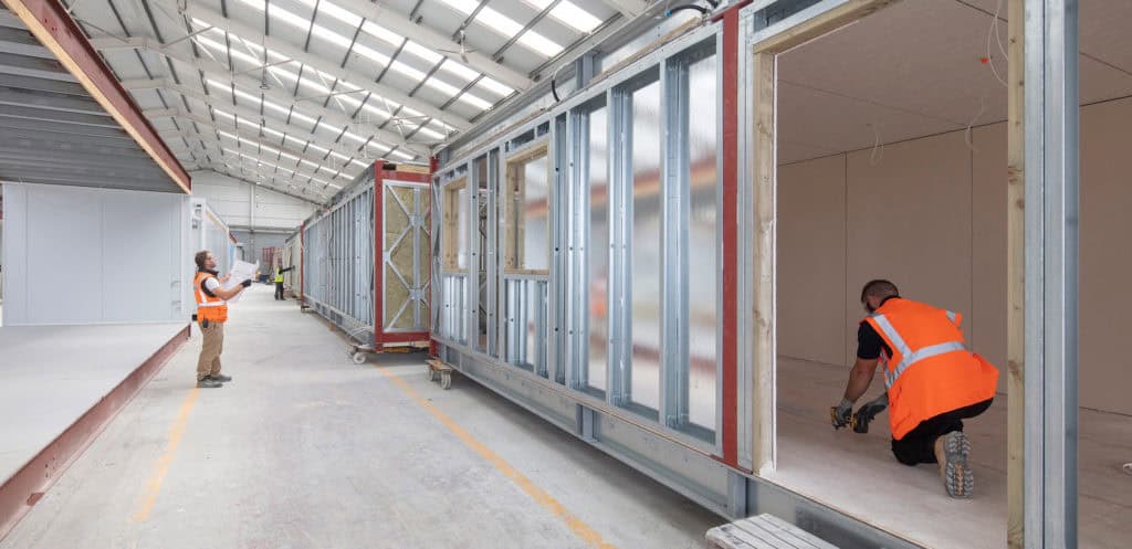 Two Premier Modular construction workers wearing orange high-vis vests, working on part of a modular building inside the offsite manufacturing site. 