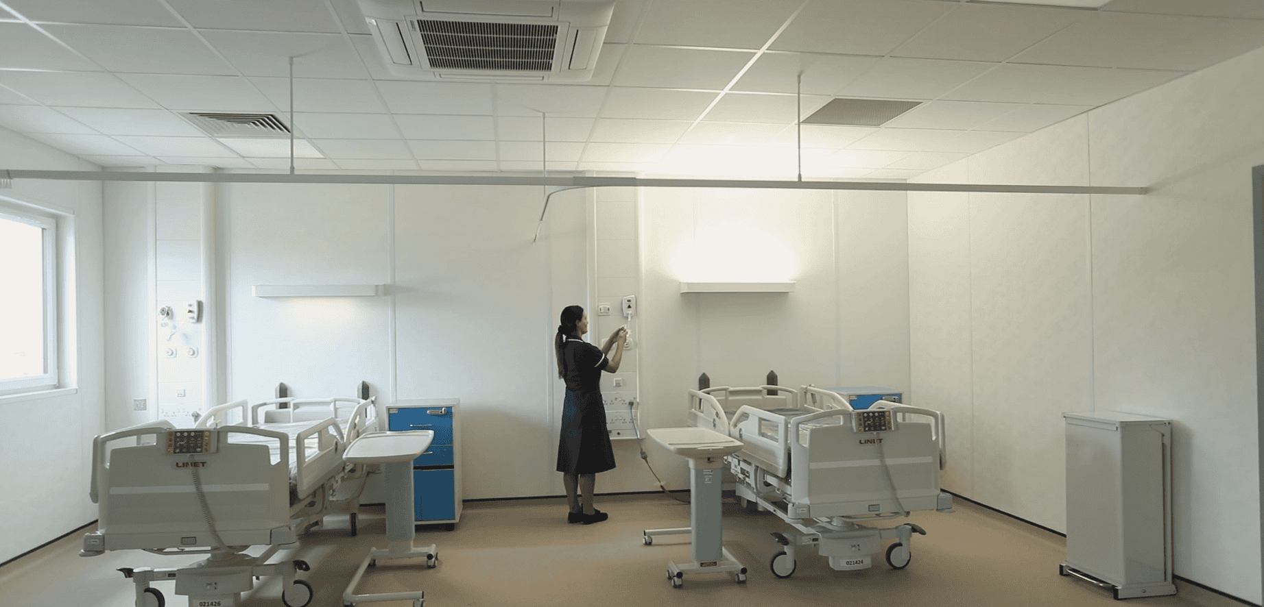 Hospital room inside a modular healthcare building. A nurse checking some machinery on the wall, two hosptial beds with desks.