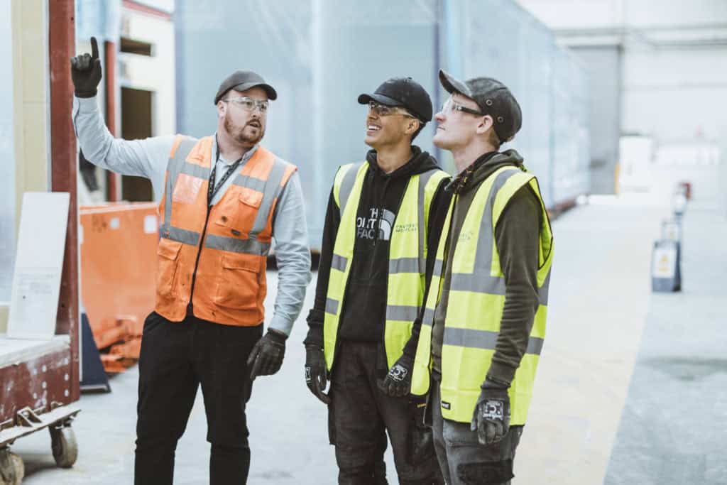 Three Premier Modular construction workers, wearing high-vis vests, caps, and protective eyewear. Stood discussing something inside the offsite construction factory.