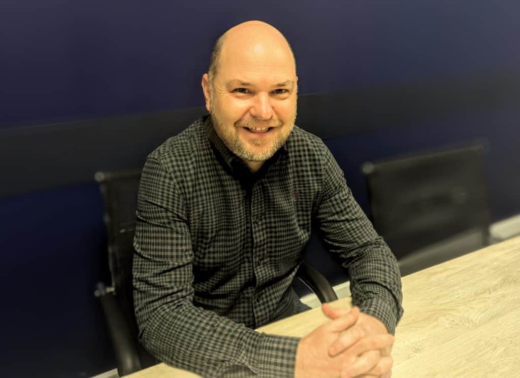 Head of Pre-Construction, Richard Hook. Bald man wearing a long sleeve shirt, sat at a desk.