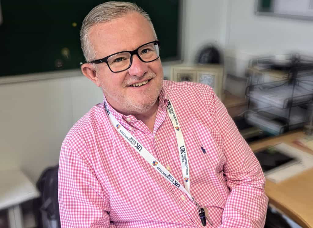 Warren Fothergill, HSEQ Manager. Man wearing glasses and a lanyard, red checkered shirt, sat at a desk.